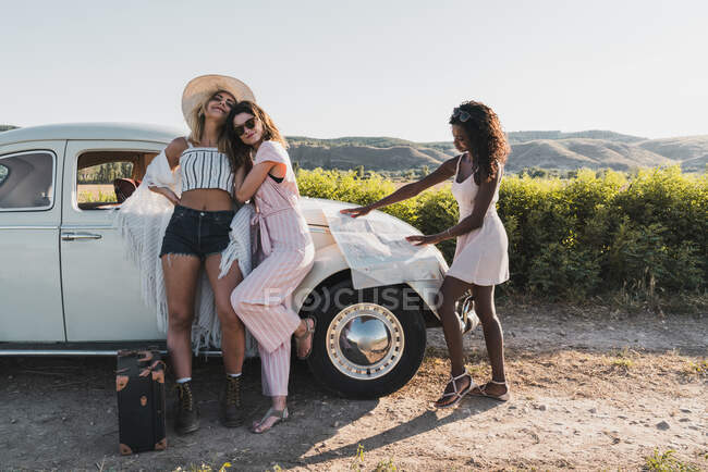 Модні багатоетнічні жінки, що стоять біля старовинного автомобіля і читають карту під час подорожей разом влітку — стокове фото