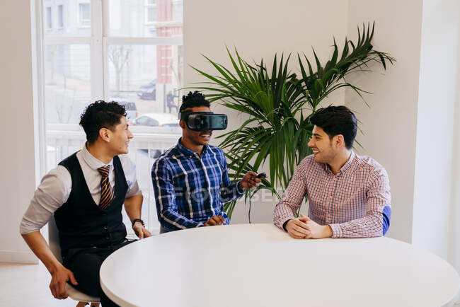 Gente alegre divirtiéndose y sentada con casco VR en la mesa de la oficina. - foto de stock