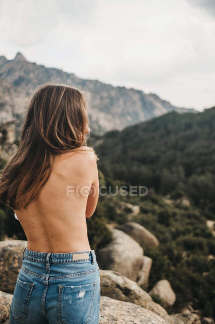 Back view of tender topless woman wearing jeans and standing on background of rocky valley in green trees — Stock Photo