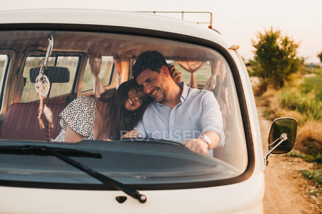 Веселый молодой человек и женщина обнимаются и едут в винтажном фургоне по дороге в сельской местности — стоковое фото
