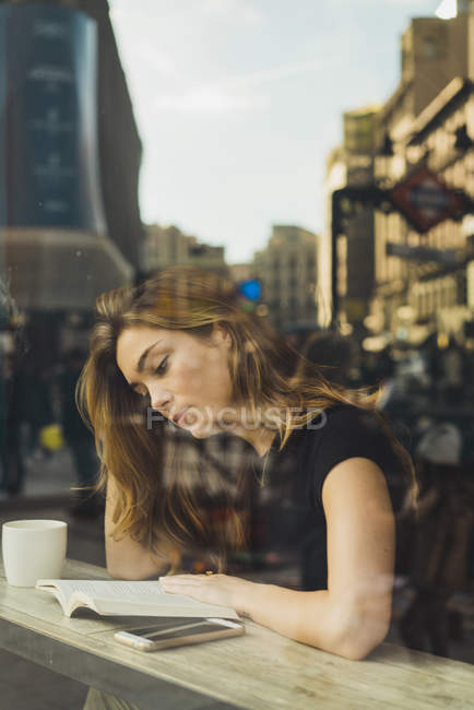 Jeune femme lisant un livre dans un café derrière une vitre avec reflet de la ville — Photo de stock