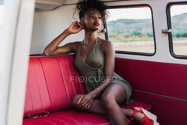 Attractive African-American female sitting on comfortable back seat of vintage van and looking away while traveling in countryside — Stock Photo
