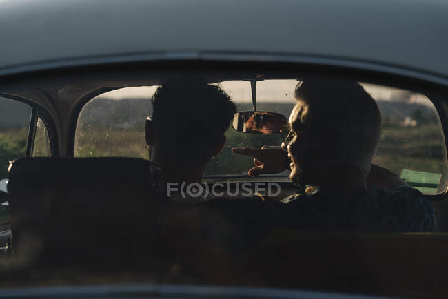 Vista trasera de dos jóvenes irreconocibles abrazando y admirando la naturaleza mientras están sentados dentro de un coche retro - foto de stock