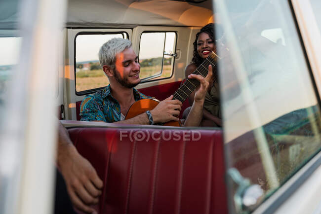 Grupo de jovens rindo e ouvindo cara bonito tocando guitarra acústica dentro de van vintage na natureza — Fotografia de Stock