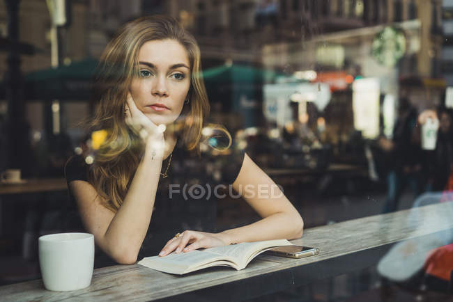 Мечтательная молодая женщина сидит с книгой и чашкой кофе в кафе — стоковое фото