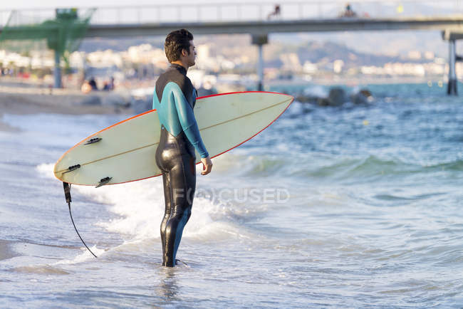 Homem com prancha de surf em pé na praia de areia molhada no oceano — Fotografia de Stock