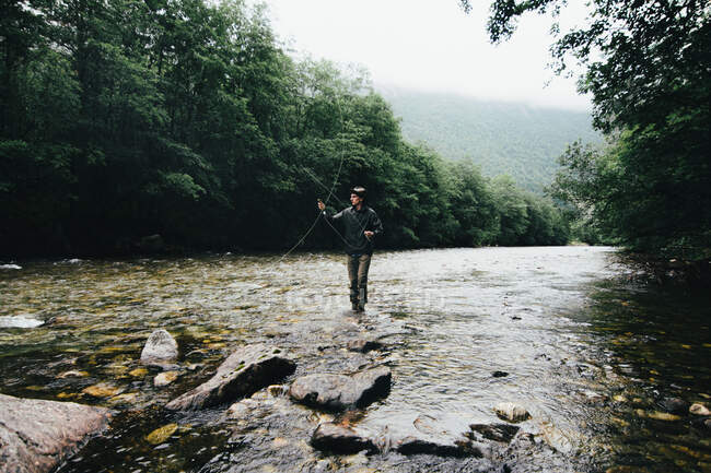 Pescador segurando vara giratória e em pé sobre rochas no fluxo do rio — Fotografia de Stock