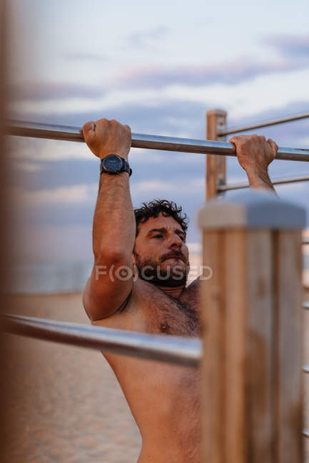 Muscular guy performing pull-ups on bar during sunset on sandy beach — Stock Photo