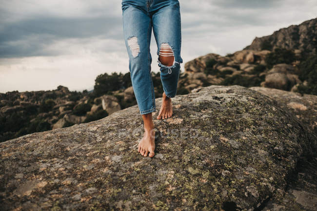 Cropped image of woman in stylish denim walking barefoot on rough gray boulder against cloudy sky — Stock Photo