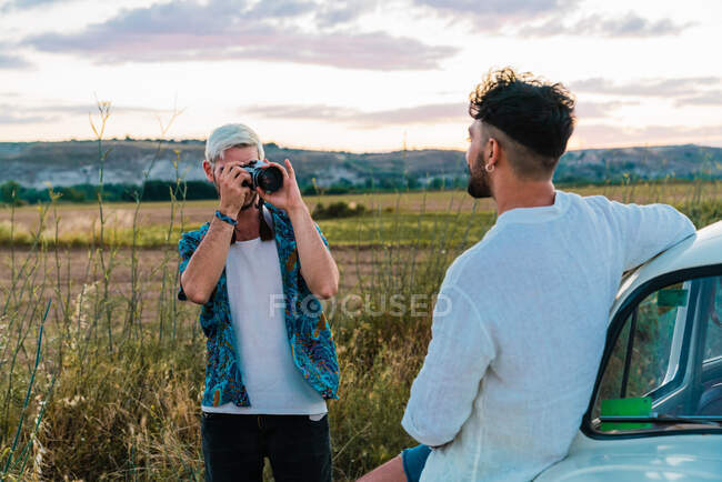 Casual homem de camisa usando câmera fotográfica e imaginando o homem com carro na paisagem do campo no verão — Fotografia de Stock