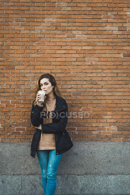 Retrato de jovem com xícara de papel de café em pé contra a parede de tijolo — Fotografia de Stock