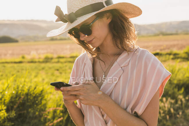 Brunette in pink clothes and elegant hat surfing smartphone on background of green summer countryside in?sunlight — Stock Photo