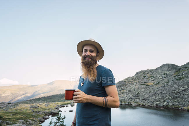 Young bearded man standing near lake in mountains with cup and looking at camera — Stock Photo