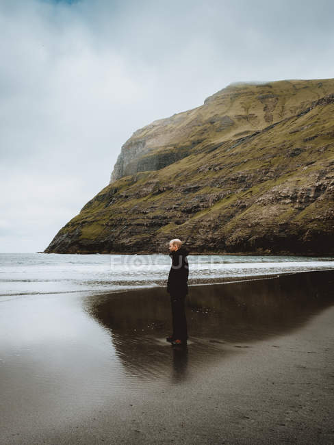 Man in warm clothes standing on shore of calm ocean with cliffs on bacground on Feroe Islands — Stock Photo