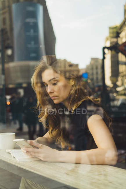 Woman using smartphone in cafe behind window pane with reflection of building — Stock Photo