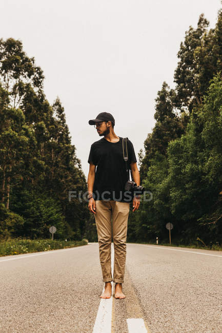 Bonito homem descalço com câmera de foto profissional olhando para longe enquanto estava no meio da estrada rural — Fotografia de Stock