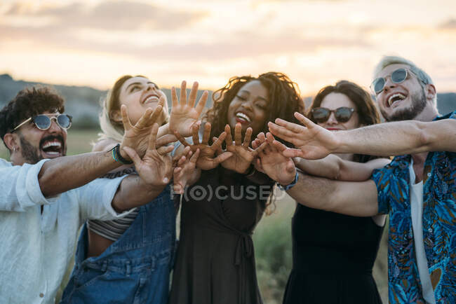 Grupo de diversos jóvenes amigos sonriendo y extendiendo las manos hacia la cámara mientras están de pie sobre un fondo borroso de un paisaje increíble durante la puesta del sol - foto de stock