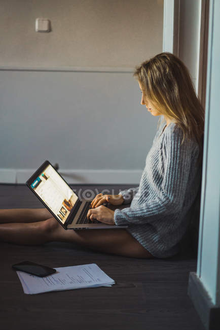 Женщина в свитере сидит на полу с документами и работает с ноутбуком — стоковое фото