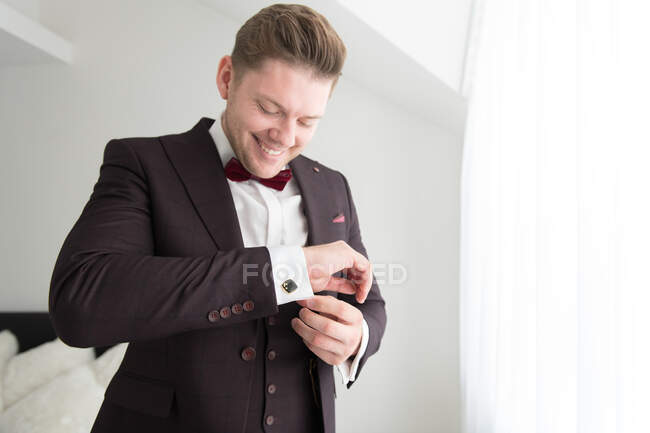 From below view of young smiling man standing in bedroom with white walls and putting on costume — Stock Photo