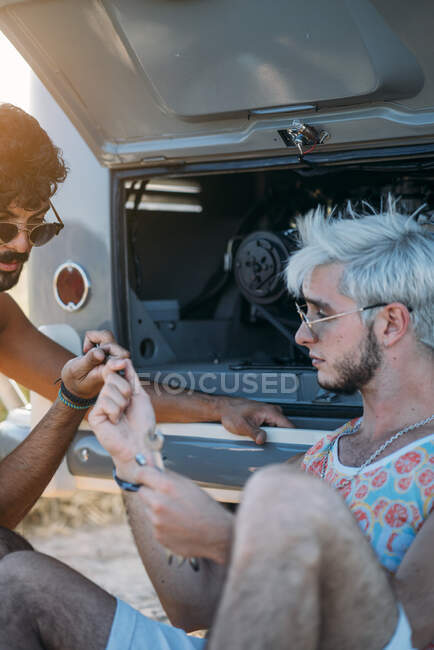 Young male leaning back and relaxing while sitting near friend trying to repair broken motor of car while?during travel in nature — Stock Photo