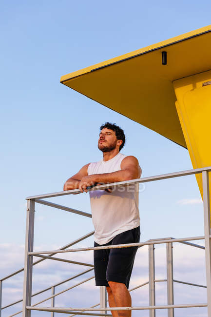 Thoughtful bearded man in sportswear leaning on railing of lifeguard cabin on beach — Stock Photo