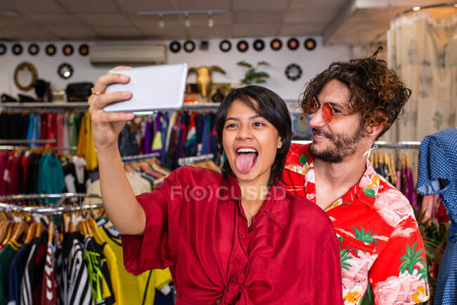 Молодий чоловік і жінка в стильному одязі посміхаються і приймають селфі, стоячи в маленькому магазині одягу — стокове фото