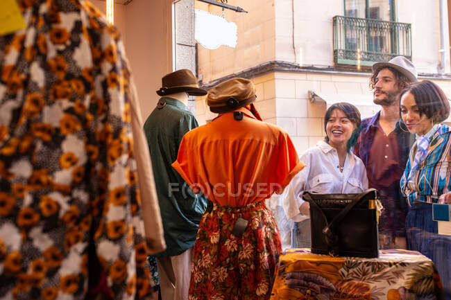 Young man and women smiling and looking at clothes inside small store while standing on street near display window — Stock Photo