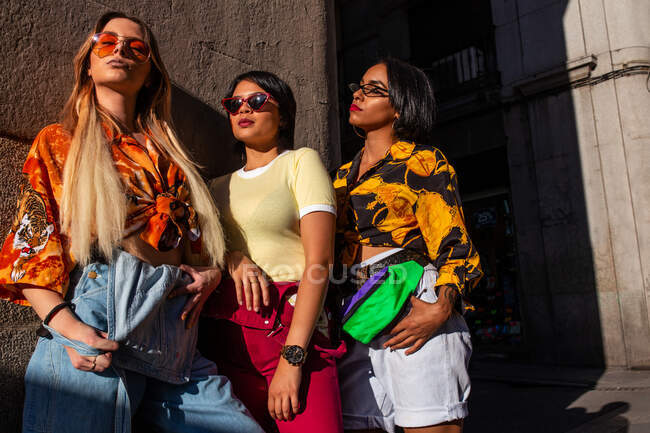 Arbre jeunes femmes dans des tenues à la mode appuyé sur le mur de construction tout en se tenant sur la rue de la ville par une journée ensoleillée — Photo de stock