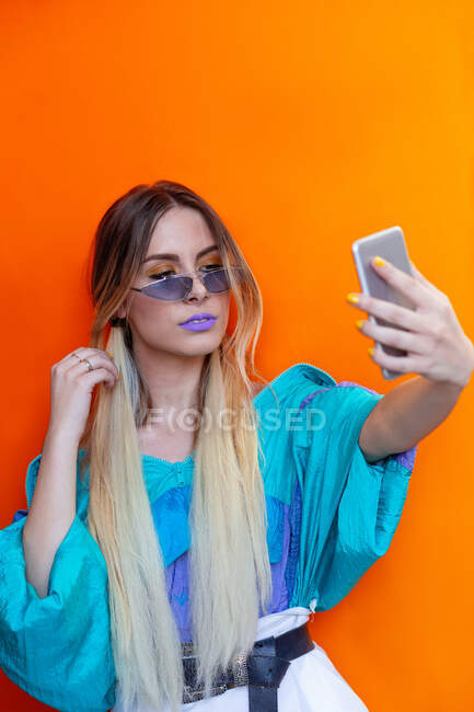 Приваблива молода жінка в модному вбранні позує за селфі, стоячи на яскраво-помаранчевому фоні — стокове фото