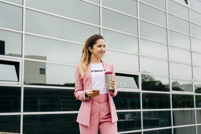 Femme gaie et tendance en costume rose avec café debout devant un immeuble de bureaux moderne — Photo de stock