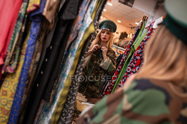 Schöne junge Frau steht in der Nähe des Spiegels und probiert Militärkleidung an, während sie Zeit in einem kleinen Geschäft verbringt — Stockfoto
