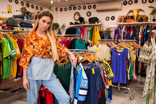 Attractive young female in trendy outfit leaning on clothes rack and looking at camera while standing in stylish shop — Stock Photo