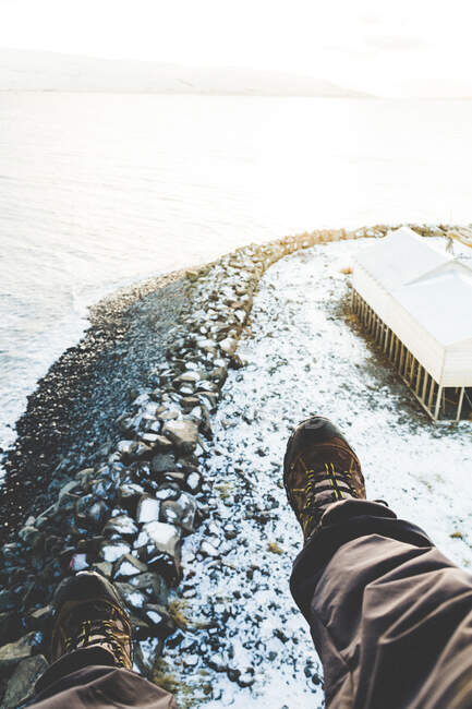 Ноги путешественника в сапогах над скалистым побережьем в снегу с палаткой и водой при ярком свете — стоковое фото