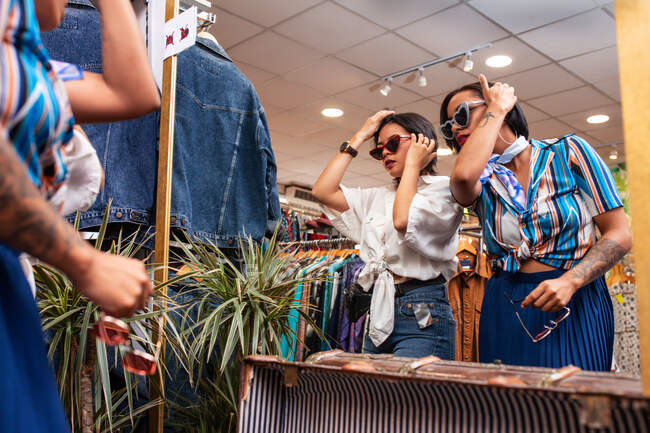 Two lovely young women in stylish outfits adjusting hair while trying on sunglasses near mirror in small clothes shop — Stock Photo