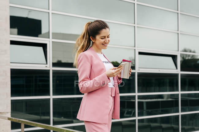Весела жінка в рожевому костюмі з кавою використовує смартфон перед сучасною офісною будівлею — стокове фото
