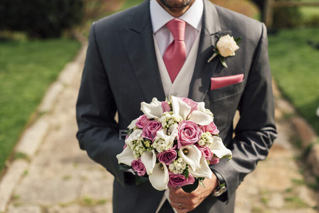Cortar hombre guapo en traje gris de pie con ramo de flores de color rosa y blanco. - foto de stock