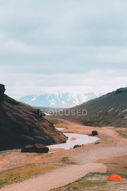 Perspective view of river in valley with mountains and small camping tent under clouds - foto de stock
