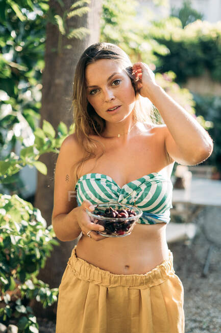 Atractiva hembra con un tazón de bayas frescas tocando el cabello y mirando a la cámara mientras está de pie sobre un fondo borroso de jardín en un día soleado - foto de stock