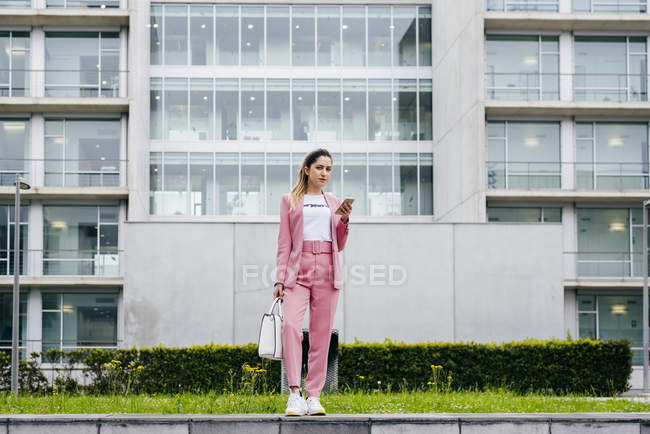 Стильна молода жінка зі смартфоном в рожевому кольорі стоїть перед сучасною офісною будівлею — стокове фото