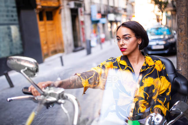 Lovely young female in trendy outfit sitting on motorcycle on blurred background of city street — Stock Photo