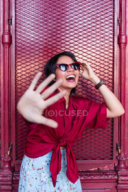 Красивая молодая женщина в модном наряде смеется и смотрит в сторону, опираясь на бордовую стену на городской улице — стоковое фото