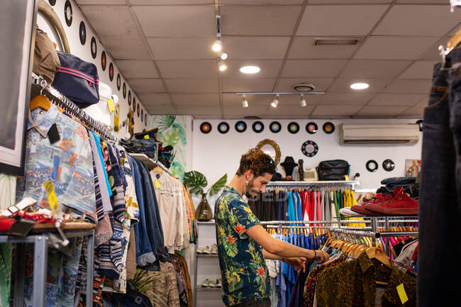 Hombre sonriendo y recogiendo prendas de vestir de carril de ropa mientras pasa tiempo en una pequeña tienda - foto de stock