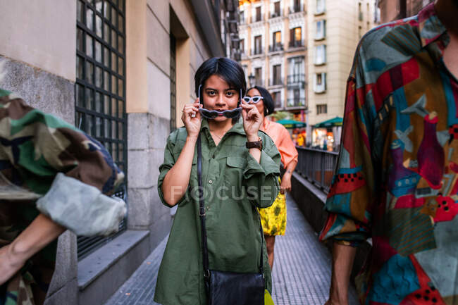 Belle femme asiatique en tenue tendance tenant des lunettes de soleil et regardant la caméra tout en marchant dans la rue de la ville parmi des amis — Photo de stock