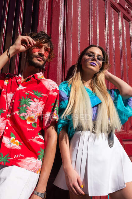 Dal basso colpo di giovane uomo e donna in abiti alla moda appoggiati a muro di colore bordeaux mentre in piedi sulla strada nella giornata di sole — Foto stock