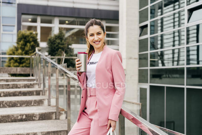 Улыбающаяся модная женщина с кофе, стоящим на ступеньках и опирающимся на металлические перила — стоковое фото