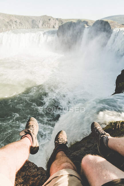 Обрізати ноги мандрівників у чоботях, що сидять на скелі над величним водоспадом у тумані та сонячному світлі — стокове фото