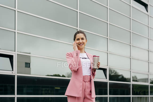 Mulher elegante alegre com café falando no smartphone em frente ao prédio de escritórios moderno — Fotografia de Stock