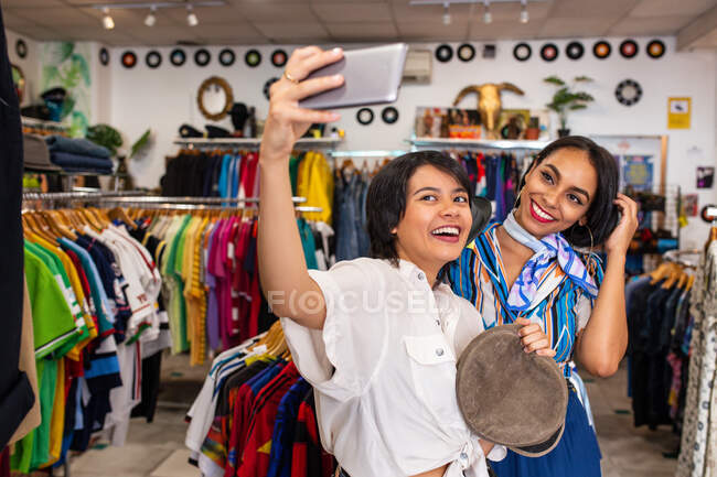 Two female friends smiling and posing for selfie while standing in small clothes shop — Stock Photo