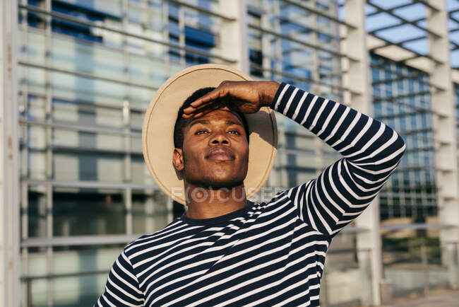 Attractive African-American man in hat keeping hand near forehead and looking at distance while standing on street — Stock Photo