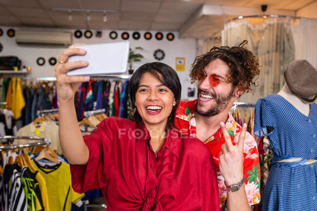 Молодой человек и женщина в стильных нарядах гримасируют и делают селфи, стоя в маленьком магазине одежды — стоковое фото